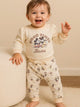 Image showing a flat lay of an Annie brand girls two-piece clothing set. The outfit includes a cream-colored long-sleeve top with a "GOOD TIMES Aloha" graphic featuring Mickey and Minnie Mouse, paired with matching cream-colored joggers covered in a whimsical all-over print of Mickey and Minnie with palm trees.