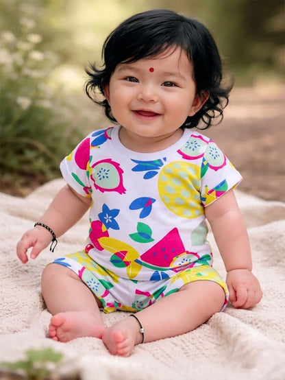 Flat lay of an Annie brand girls two-piece clothing set featuring a white short-sleeve t-shirt and matching shorts with a vibrant all-over tropical fruit print including watermelon pineapple and dragon fruit. The shorts have an elastic waist with a white decorative tie front pockets and cuffed hems.