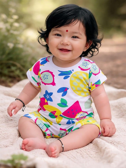 Flat lay of an Annie brand girls two-piece clothing set featuring a white short-sleeve t-shirt and matching shorts with a vibrant all-over tropical fruit print including watermelon pineapple and dragon fruit. The shorts have an elastic waist with a white decorative tie front pockets and cuffed hems.