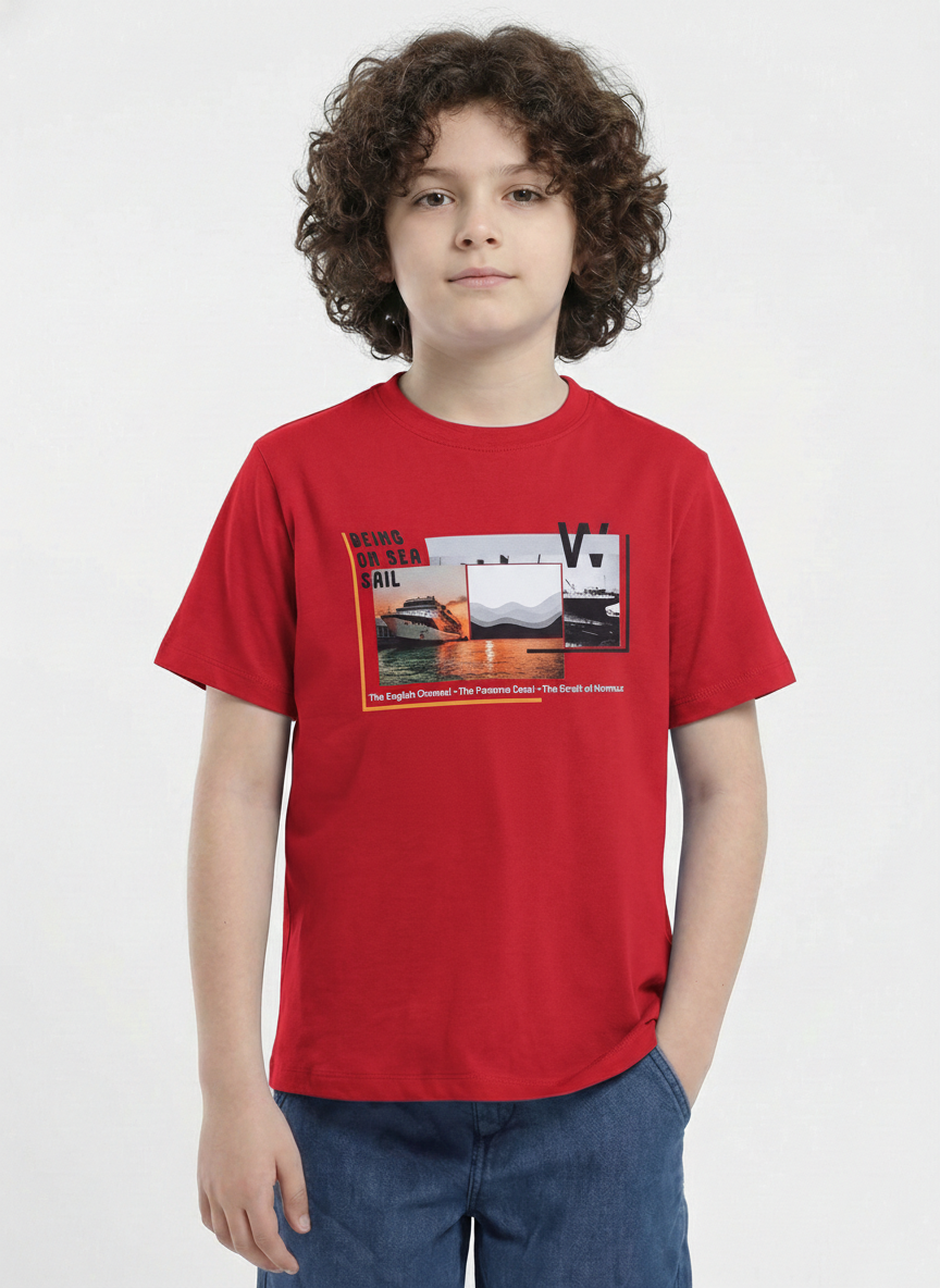 showing a young boy with curly hair modeling a Biskid brand bright red short-sleeve t-shirt. The shirt features a rectangular chest graphic with a sunset, a cruise ship, and the text "BEING ON SEA SAIL" along with famous global waterways.