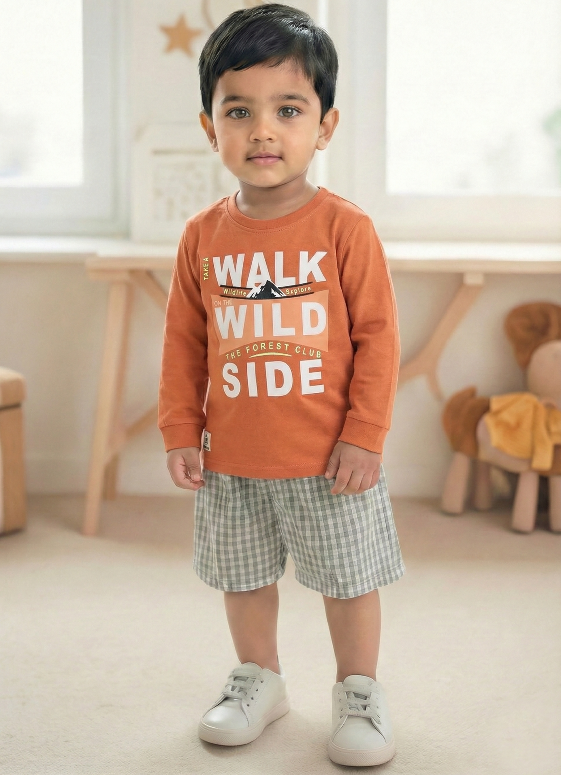 Image file Gemini_Generated_Image_pobj3hpobj3hpobj.jpg showing a young boy wearing a rust-orange Biskid brand boys long-sleeve t-shirt with a "Walk on the Wild Side" forest graphic on the chest, paired with gray checkered shorts and white sneakers.
