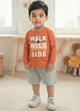 Image file Gemini_Generated_Image_pobj3hpobj3hpobj.jpg showing a young boy wearing a rust-orange Biskid brand boys long-sleeve t-shirt with a "Walk on the Wild Side" forest graphic on the chest, paired with gray checkered shorts and white sneakers.