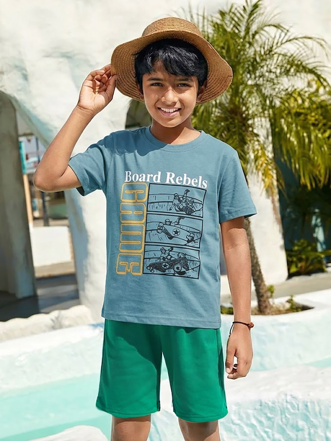 Image file 877-P.Blue.jpg showing a Biskid brand boys' powder blue crew neck t-shirt with a "Board Rebels" comic-style skater graphic on the front, displayed on a wooden hanger with a toy camera and colorful "GAME" letter blocks nearby.