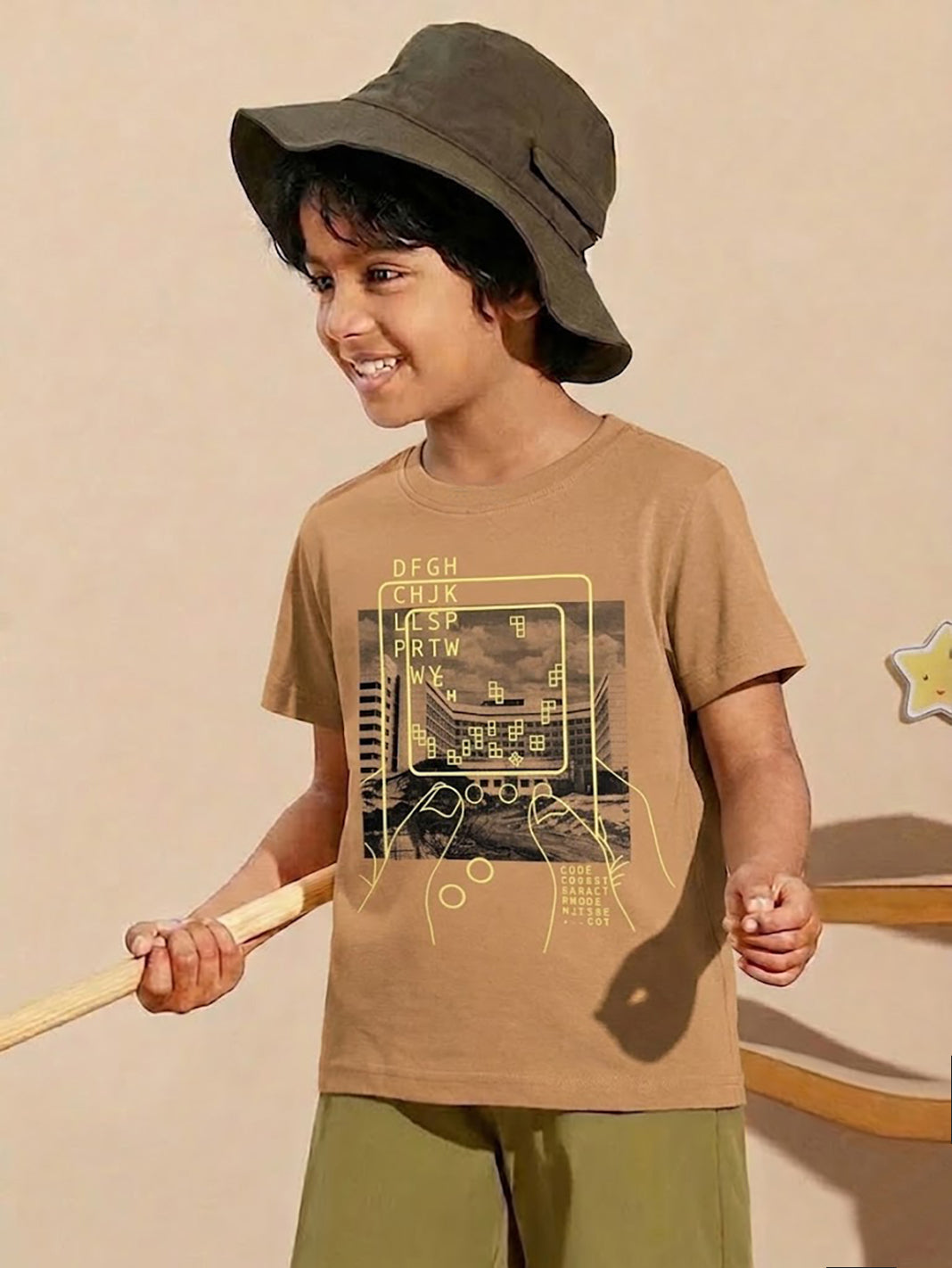 Image file 877-Beige.jpg showing a Biskid brand boys' beige crew neck t-shirt with a retro gaming console graphic on the front, displayed on a wooden hanger with dice and star-shaped decorations nearby.