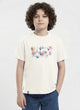 a young boy with curly hair modeling a Biskid brand cream-colored short-sleeve t-shirt. The shirt features a geometric graphic made of colorful pastel hexagons and triangles containing travel photos, accompanied by the text "TO TRAVEL IS TO LIVE".